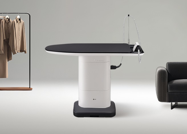 회색배경에 좌측에는 베이지색 의류들이 헹거에 걸려있고 중앙에 시스템 아이어닝 제품이 있습니다. 우측에 검은색 소파가 배치되어있습니다.​